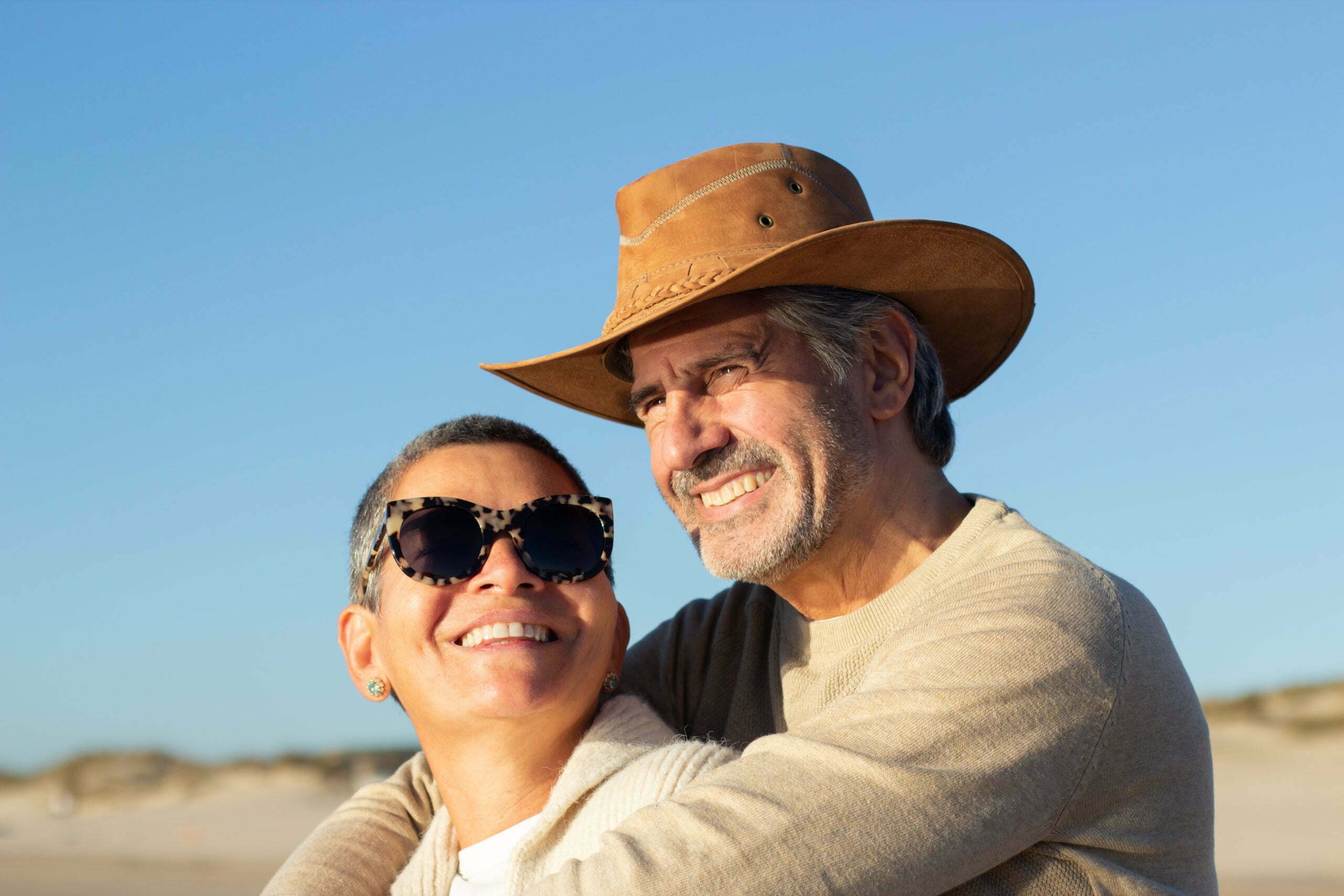 Portrait of beautiful senior couple spending time together outdoors on sunny day. Handsome smiling grey-haired man in cowboy hat hugging happy short-haired lady in sunglasses. Love, leisure concept
